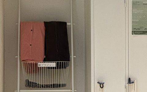 更衣室。白いハシゴ型の棚にピンクと黒の館内着がかかり、「紙ブラジャー」の札がついたカゴと、ロッカーの扉が見える