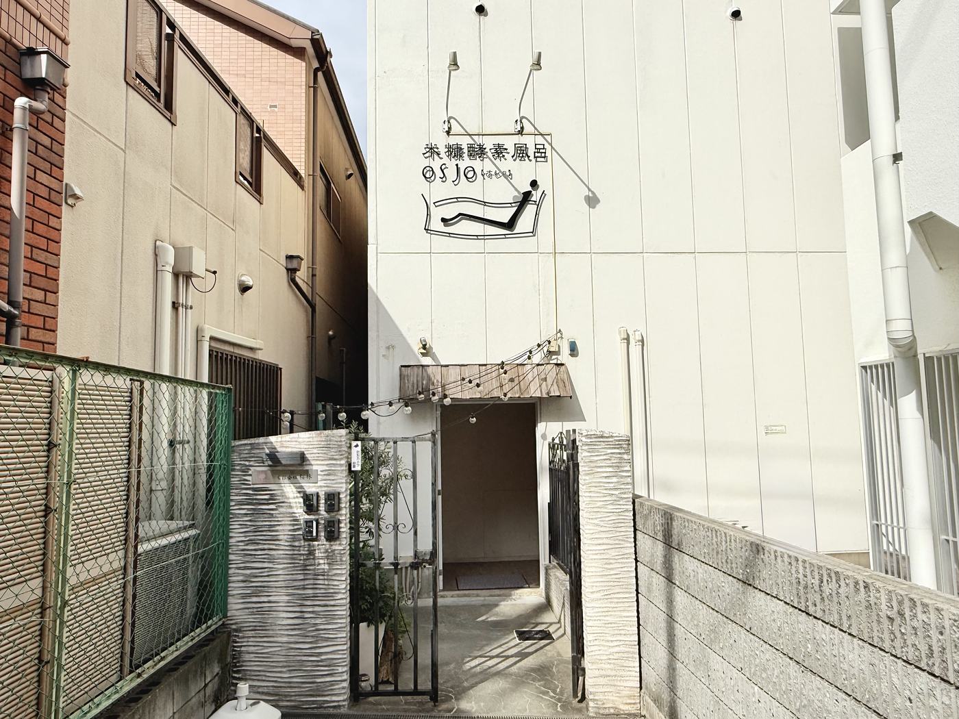 建物の外観。白い壁に osjo のロゴの看板があり、門の奥が入口