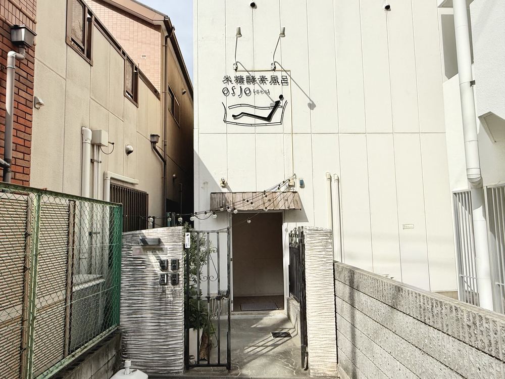建物の外観。白い壁に osjo のロゴの看板があり、門の奥が入口